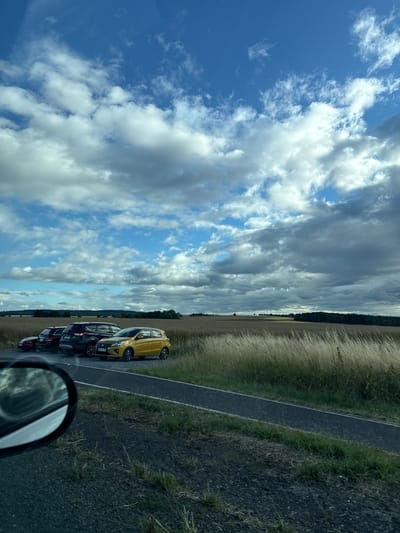 The image depicts a parking area situated in the rural surroundings of Weidenhausen, Germany. The scene features a row of parked vehicles, including a red hatchback, a black sedan, a black SUV, and a yellow hatchback. The cars are parked along a paved surface adjacent to a field of tall, dry grass and crops. A winding road is visible in the foreground, with a car's side mirror reflecting a portion of the sky and surrounding landscape. The sky above is characterized by a mix of blue patches and dramatic, heavy clouds. The overall environment suggests a rural landscape, with fields stretching towards a treeline in the distance.