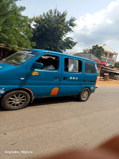 A blue van with the license plate C-B-7 is driving on a paved road in the city of Okinni, Nigeria, located at coordinates 7.832 latitude and 4.51 longitude. The van is traveling from left to right in the image. There is a tree on the left side of the image and a building on the right side of the image. The building appears to be a bank, with the word "BANK" visible on the exterior. It is a sunny day with a few clouds in the sky. The van is traveling at a moderate speed, and there are no other vehicles on the road. There are no pedestrians visible in the image. The van is the primary subject of the image.