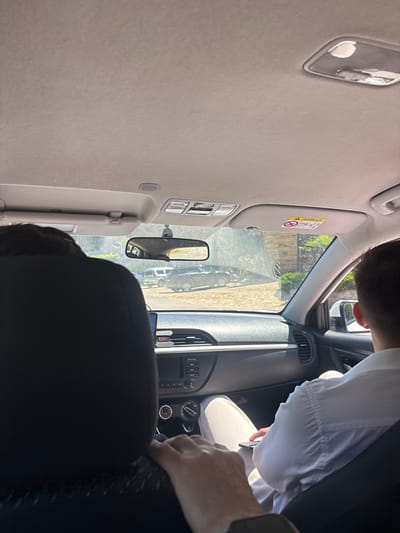 The image shows the interior of a car, looking forward from the back seat. In the foreground, the dark fabric of the back of the driver's seat is visible on the left. A person's arm and hand are resting on the headrest of the seat in front. To the right, the back of a person wearing a white shirt and light-colored pants is visible, and this person appears to be holding a dark-colored object, possibly a phone. The car's dashboard is visible, including the infotainment system and climate controls. A rear-view mirror is mounted on the windshield, reflecting an outdoor scene with cars parked. A sun visor with an airbag warning label is also visible. The environment outside the car appears to be sunny and possibly rural or suburban, with greenery and buildings visible in the distance. The photo was taken in Kazachiy Brod, Russia.