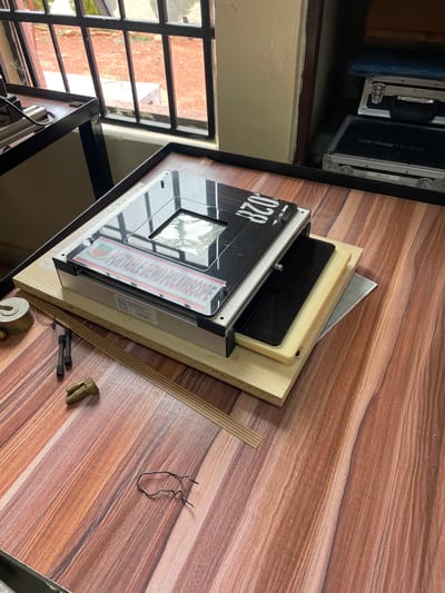 The image depicts a cluttered scene on a wood-grain surface. A "Portable Demo Polariscope" marked with "028" sits atop stacked pieces of wood and graph paper. Scattered around the polariscope are small metallic objects, including what appear to be screws and a wire clip. A portion of a metal-framed window with bars is visible in the background, suggesting an interior location. Based on additional information, the location is Afaka, Nigeria.