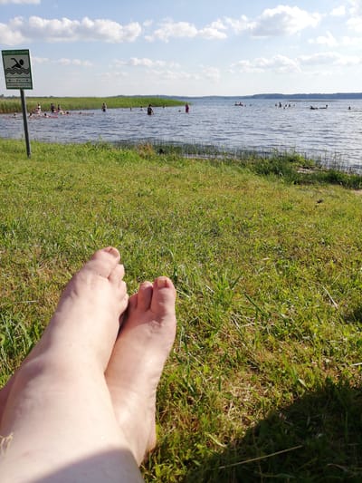 The media file depicts a sunny day at a lake or river with a grassy embankment in the foreground. The camera's perspective is low, focusing on a pair of bare feet resting on the grass. In the background, a body of water stretches towards a treeline on the horizon. Several people are visible in the water, some swimming and others wading. A signpost stands on the left side of the image, near the water's edge. The sign has text in Cyrillic, which reads "МЕСТО ДЛЯ КУПАНИЯ" (Place for swimming) and indicates a distance of "40 м" (40 m). The weather appears to be clear with a few white clouds in a blue sky, suggesting it is daytime. The overall mood is relaxed and summery, indicating a recreational outing. The location context is identified as Peryabrodski Sel'ski Sovet, Belarus.