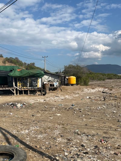 In the city of Upt Tambak Sari, Indonesia, a scene of disarray unfolds under a bright sky dotted with white clouds. The foreground is dominated by a large expanse of dry, barren land littered with a significant amount of debris. This includes plastic bottles, wrappers, discarded tires, and various other unidentifiable refuse, creating a landscape of neglect.

In the mid-ground, a makeshift structure stands, appearing to be a small shop or dwelling. It is constructed from salvaged materials, including corrugated metal and wooden planks. A green tarp is draped over a portion of the structure, and a large, bright orange water tank is visible nearby, resting on tires. A discarded tire is also present in front of the structure, adding to the cluttered environment. The structure itself shows signs of wear and tear, with peeling paint and exposed wooden supports.

Further in the background, a lush green hillside rises, contrasting with the desolate foreground. Beyond the hillside, a large, dark mountain looms under the partly cloudy sky. Power lines crisscross the scene, suspended from utility poles, indicating the presence of infrastructure amidst the otherwise unkempt surroundings. The overall impression is one of a neglected and impoverished area, characterized by widespread litter and makeshift constructions.