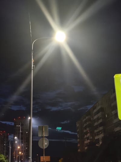 An eye-level, wide-angle shot captures a streetlamp illuminating a dark, cloudy night sky over a city. The streetlamp, positioned in the left half of the frame, emits a bright, starburst-like light, with beams extending outwards and creating lens flares. Below the streetlamp, a traffic sign with two circular signs is visible. To the right of the streetlamp, a building with multiple balconies and windows is partially lit by the lamp. In the background, other buildings with illuminated windows are visible, along with streetlights. The clouds in the sky are dark and appear textured, suggesting movement. The overall scene is illuminated by the streetlamp and ambient city lights, creating a moody and somewhat dramatic atmosphere.