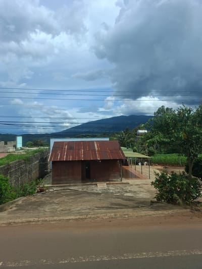 The image depicts a rural scene in Tân Phú 2, Vietnam, featuring a small, weathered, tin-roofed house as the central subject. The house appears to be constructed with dark wooden planks and has a single entrance. Adjacent to the house, a covered patio area with a tiled floor is visible, supported by metal poles. Beyond the immediate structures, a concrete wall with overgrown vegetation lines the left side of the frame. In the background, a body of water or low-lying land is visible, leading to a range of distant mountains under a cloudy sky. Power lines crisscross the upper portion of the image, obscuring parts of the sky and mountains. A large green tree with red flowers stands to the right, partially obscuring the view of the surrounding fields and a water tower with a silver tank. The foreground shows a paved surface, possibly a road, with white markings. The overall impression is of a simple, somewhat isolated dwelling in a natural, possibly agricultural, setting.