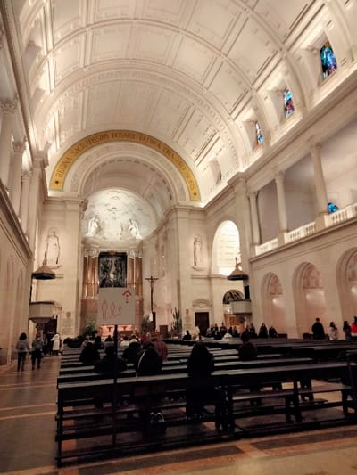 This image offers a wide-angle view of the majestic interior of a grand basilica, likely the Basilica of Our Lady of the Rosary in Fátima, Portugal, as indicated by the prominent inscription. The scene is dominated by impressive classical architecture, featuring a soaring vaulted ceiling with intricate coffering and decorative panels. Tall, ornate columns line the nave, supporting a series of arches and upper galleries.
In the foreground and midground, numerous dark wooden pews are occupied by visitors or congregants, many facing towards the front of the church. They appear to be engaged in prayer, quiet contemplation, or attending a service, creating a serene and reverent atmosphere. A white sign with red pictograms of people and an arrow pointing up is visible in the aisle, potentially guiding movement or indicating social distancing measures.
At the far end, the main altar area is illuminated, showcasing a large, ornate altarpiece with religious sculptures or a mosaic, flanked by statues of saints. A crucifix stands to the right of the altar. High on the right wall, several small, colorful stained-glass windows add subtle hues to the vast interior.
A key detail is the golden inscription arching gracefully above the altar: "REGINA SACRATISSIMI ROSARII FATIMA ORA PRO NOBIS." This Latin phrase translates to "Queen of the Most Holy Rosary, Fátima, pray for us," explicitly confirming the sacred location. The entire space is brightly lit by artificial light sources, enhancing the grandeur of the architecture and the solemnity of the setting. The image captures a moment of quiet devotion within this magnificent religious edifice.