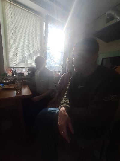 An indoor scene in Bolshaya Elhovka, Russia, depicts three individuals seated around a wooden table, illuminated by strong sunlight entering through a large window on the left. The window is partially covered by white horizontal Venetian blinds, and bare tree branches are visible outside. The intense backlighting from the window causes significant lens flare and renders the individuals in various states of silhouette or underexposure.
On the far left, a person is seated, wearing a light-colored short-sleeved top, with their face obscured by light. To their right, a second individual is seated, wearing a patterned long-sleeved top, also with their face largely obscured. In the foreground on the right, a third person is seated, wearing a dark-colored long-sleeved top, possibly with a Nike logo visible on the chest, and blue trousers. This individual's left hand rests on their thigh, displaying a ring on the fourth finger.
On the wooden table, a clear glass containing an amber-colored liquid is present, along with a light-colored dish. The windowsill beneath the blinds holds various small, indistinct objects. The ceiling in the upper right features a silver-colored corrugated ventilation duct. The room's general appearance suggests a residential setting, with the background on the right side being dark and indistinct, showing a potential screen or television. The overall atmosphere suggests a casual gathering or meal.