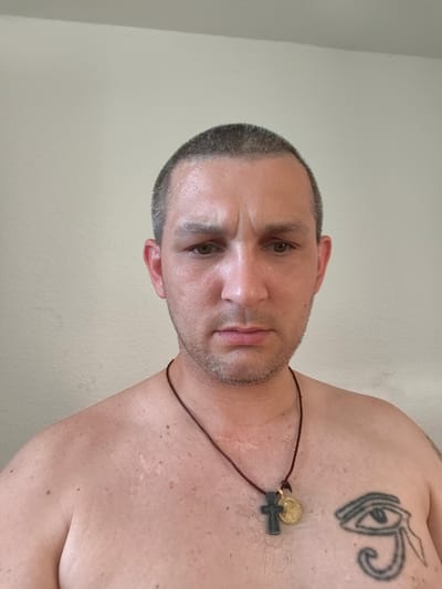 A man with short, dark hair is looking directly at the camera. He is shirtless, and wearing a necklace with two pendants: a dark cross and a gold circular medal. He has a tattoo on his chest that resembles the Eye of Horus. The background is a plain, light-colored wall. The image is a head and torso shot, taken from a medium distance. There is no identifiable location in the city of Wolgast, Germany visible in the image.