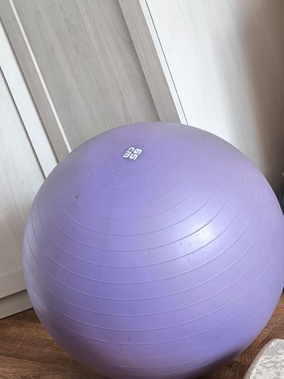 A large, purple exercise ball is centered in the frame, resting on a wooden floor. The ball has textured concentric rings and a white logo with the letters "SG" is visible on its surface. Behind the ball, a light-colored wall with vertical paneling is visible. To the right of the ball, a dark object, possibly another piece of furniture or equipment, is partially visible. The overall scene suggests an indoor setting, possibly a home gym or a personal space. The location in Gal’chino, Russia is not visually discernible from the image.