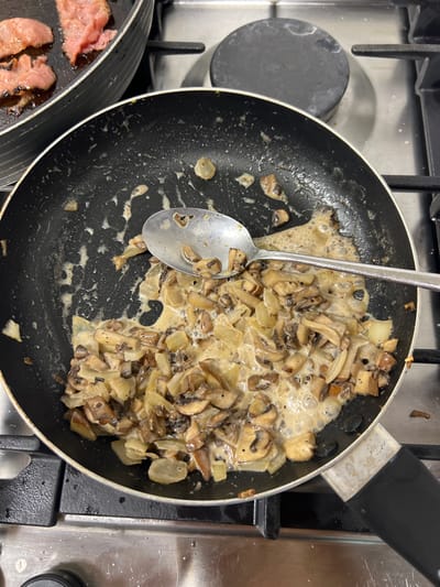 The image depicts a detailed, top-down view of an ongoing food preparation scene within a kitchen in Hofit, Israel. Two black non-stick frying pans are positioned on a stainless steel gas stovetop equipped with black grates. The prominent pan in the foreground contains a substantial quantity of sautéed, sliced mushrooms and diced onions immersed in a light-colored, possibly creamy or buttery sauce. A silver metal spoon rests diagonally within this mixture, partially submerged. This pan shows internal residue and wear. In the upper-left background, a second, partially visible black non-stick frying pan holds several pieces of cooked, rendered meat, consistent with bacon or similar cured pork products. The gas stovetop surface exhibits splatters and general residue from cooking activity around the burners. No individuals are present in the frame.