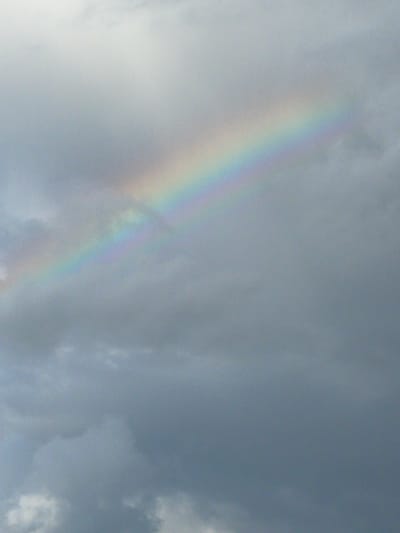The image depicts a sky filled with dark, overcast clouds, with a faint rainbow arching across the upper right portion of the frame. The rainbow displays a spectrum of colors, including red, orange, yellow, green, blue, and violet, although they are somewhat muted due to the cloudy conditions. The lower part of the image is dominated by the dense gray clouds, suggesting a recent or impending rainfall. There are no people, structures, or specific landmarks visible that would indicate the location within Zaķumuiža, Latvia. The environment is solely characterized by the cloudy sky and the presence of the rainbow.