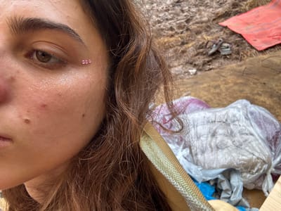 A close-up, slightly rotated, photo shows a woman’s face with brown hair. She has glitter on her forehead and three small pink gems above her eye. Her complexion is fair. In the background, there is a bag of crumpled plastic, a wooden surface, and a small red cloth. The photo appears to be taken indoors, with a focus on the woman's face and artistic makeup.