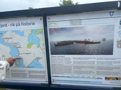 This media file features a photograph of an outdoor information board detailing the Battle of Hafrsfjord, a significant event in Norwegian history. The board is divided into sections, with a map on the left and text and images on the right.
The map displays a fjord system with several points of interest marked, including "Helleristninger Gravrøyser" (Rock Art Burial Cairns), "Naust Boathouse," and "Fluberget Helleristninger" (Rock Art). A red line indicates a footpath, and a person's hand is pointing to a section of the path near the water.
The right side of the board features an illustration of a fleet of Viking longships on the water, with a large body of men on board each vessel. The text provides historical information about the Battle of Hafrsfjord, its significance in the unification of Norway under King Harald Fairhair, and the monument "Sverd i fjell" (Sword in the Rock) commemorating the event. It also mentions archaeological finds and the historical use of the Hafrsfjord area during the Iron Age.
Visible text includes "fjord - rik på historie" (fjord - rich in history), "Slaget i Hafrsfjord" (The Battle of Hafrsfjord), "The Battle at Hafrsfjord," and various Norwegian historical periods such as "Eldre steinalder" (Old Stone Age) and "Vikingtid" (Viking Age), along with their corresponding date ranges. The name "Rogaland Fylkeskommune" is also visible, indicating the regional government's involvement in the information provided. The overall scene suggests a historical site with an educational display, likely in a public outdoor setting given the sunlight and natural background elements.