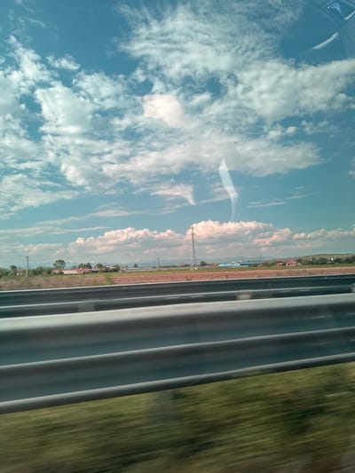 A blurred, mobile view from a train window shows a landscape in Shishmantsi, Bulgaria. To the left of the frame, a clear blue sky is filled with scattered white clouds. Reflected in the window is an overhead view from inside the train, as well as a village landscape that includes fields, trees, and some buildings, including those painted red. There's also a thin, possibly dusty layer of earth in between the village and the fields. Rightmost are gray fabric curtains that give way to a blur of green vegetation and brown earth, indicating rapid movement. The focus is primarily on the village outside the window and the curtain that blocks the view, but the lack of clear features makes it difficult to distinguish details.