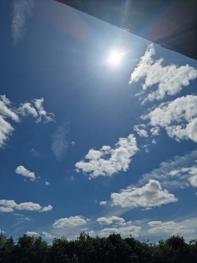 A bright, sunny day over Fenland District, United Kingdom. The sky is a vibrant blue, dotted with fluffy white cumulus clouds. The sun, a brilliant orb, shines intensely in the upper left quadrant of the frame, casting rays across the sky. A dark, horizontal structure, possibly an overhang or a building edge, is visible at the very top of the image. Below the sky, a dense line of dark green trees forms the horizon. Thin, wispy clouds streak across the sky, adding depth and texture. The scene suggests a clear and warm day, with good visibility.