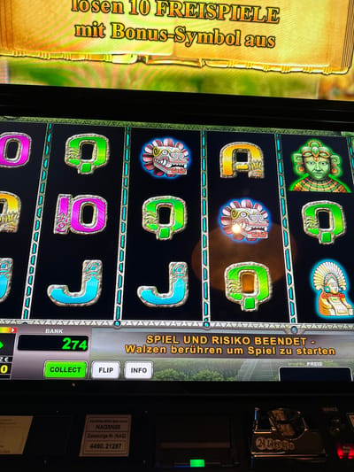 The image displays a close-up, angled view of a digital slot machine screen within what appears to be an arcade or gambling establishment in Lohfelden, Germany.

The screen exhibits a game interface with a dark background. At the top, a gold-colored banner with a textured background displays the German text "losen 10 FREISPIELE mit Bonus-Symbol aus" (win 10 FREE SPINS with Bonus symbol). Below this banner are multiple vertical reels featuring various symbols. Visible symbols include stylized letters O (pink), Q (green), J (light blue), and 10 (pink), alongside Aztec/Mayan-themed graphics such as a serpent head (red, white, blue), a green-skinned man's face with a headdress, and a seated figure with a large white headdress. The serpent head symbol in the middle column, middle row, and top row appears illuminated.

At the bottom of the screen, a control panel interface is visible. A box labeled "BANK" displays "274" in green text. Three interactive buttons are present: a green "COLLECT" button, a "FLIP" button, and an "INFO" button. To the right, a message in German states: "SPIEL UND RISIKO BEENDET - Walzen berühren um Spiel zu starten" (GAME AND RISK ENDED - Touch reels to start game).

Below the digital screen, on the physical machine's control panel, a white label with black text is visible, partially obscured. Identifiable text includes "SPIELGERÄT (Btm GewQ)", "NAG5N08", and "Zulassungs-Nr. (N.A.G) 4490.21287". A small, rectangular green light is illuminated on the panel to the left of a partial view of a coin slot or currency acceptor mechanism. The overall environment appears dimly lit, contrasting with the bright screen. No individuals are visible in the frame.