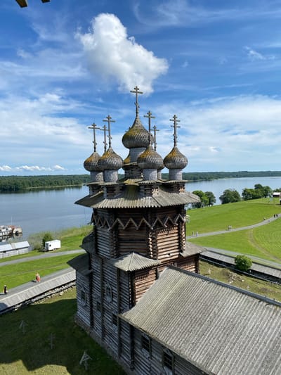 An aerial, eye-level, medium shot captures the wooden Transfiguration Church on Kizhi Island, Boyarshchina, Russia. The church, with its multiple onion domes topped with crosses, is the central subject. The domes are weathered and grey, adorned with traditional wooden shingles. The church's walls are constructed from logs, with visible horizontal timber stacking. Small, multi-paned windows punctuate the wooden facade. A sloping, grey, shingled roof is visible in the foreground.
The church is situated on a grassy knoll, with a pathway curving around it. Several small figures are seen walking on this path, enjoying the outdoor environment. The pathway is lined with wooden fencing. Beyond the church and the grassy area, a wide, calm body of water, likely Lake Onega, stretches to the horizon. A dense line of green trees borders the far side of the lake. The sky above is a vibrant blue, populated by scattered white clouds, one large fluffy cloud being particularly prominent. In the background, a small white trailer is parked near the water's edge, and a pier with a docked boat is also visible. The overall scene conveys a sense of historical architecture set within a natural, tranquil landscape.