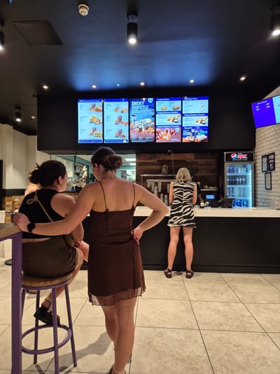 Here is a description of the image:
The image shows the interior of a Taco Bell restaurant. Two women are seated at a table in the foreground. One woman is wearing a brown dress and the other is wearing a black top and green skirt. A third woman in a zebra print dress is standing at the order counter in the background. The menu boards display various food items and prices. Drinks are visible in a refrigerated display case.