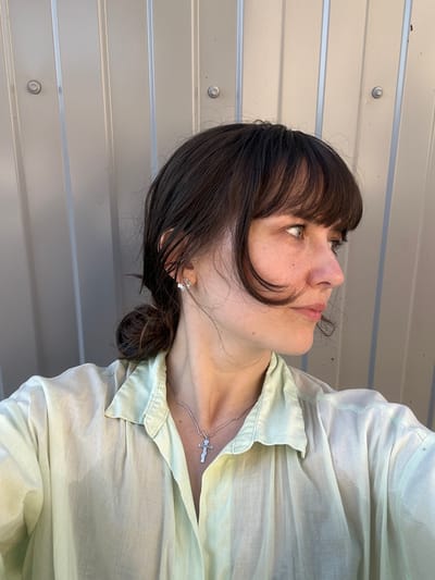 A young woman with dark hair pulled back in a low bun, bangs falling around her face, is captured in a close-up profile shot against a corrugated metal wall. She wears a pale green, oversized collared shirt and a silver necklace with a cross pendant. Her head is turned to the right, with her gaze directed off-camera. The setting appears to be outdoors, with the metallic sheen of the wall suggesting an industrial or utilitarian structure. The natural lighting indicates it is daytime. The woman has light skin, a smattering of freckles, and is wearing small, stud earrings.