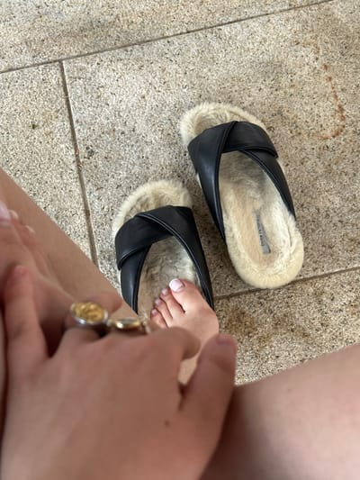 The scene is set on a light-colored, textured tiled surface within the city of Vandœuvres, Switzerland; the exact location within the city is not discernible. The image focuses on a person's lower extremities and two slippers. The person's right foot is partially inserted into the left slipper, revealing five toes with white-tipped nails. A left hand is visible in the foreground, wearing two rings: one silver-toned with a circular gold-toned emblem, and another gold-toned band. Two slippers, featuring black leather crossover straps and a cream-colored faux fur lining and footbed, are present. The right slipper's insole displays the brand name "Emme Parsons." The slippers rest on square-shaped, light-colored floor tiles with visible grout lines. No other individuals or significant objects are visible.