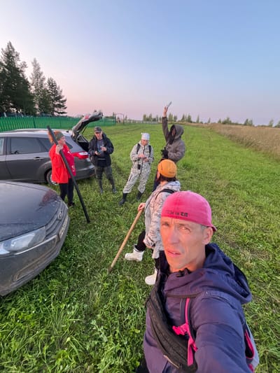 The image depicts a group of six individuals in an outdoor, grassy environment with two cars parked nearby. The scene appears to be located near the village of Derevnya Perebor, Russia, suggested by the overall setting of rural landscape and the presence of cars, implying a potential outing or activity.
In the foreground, a man wearing a pink baseball cap and a blue hooded jacket is taking a selfie, holding the camera with his left hand. He has a black fanny pack worn across his chest. His cap has text on it that reads "I'LL BE IN LA."
Behind him, to his right, a person in a camouflage-patterned outfit and a yellow beanie is holding a wooden pole. To the left of this individual, another person in a similar camouflage outfit and a white bandana is looking at their phone, which is held in their left hand.
Further back and to the left, a woman in a bright orange jacket and a patterned headband is holding a long, dark pole vertically with both hands. She is standing next to the open rear of a dark gray car.
Behind the woman in orange, a man in a dark jacket and a cap is standing near the open trunk of the gray car, holding a phone in his right hand.
To the right of the man in the dark jacket, a person wearing a dark hooded jacket with the hood up is holding a phone or similar object aloft in their right hand, seemingly pointing it towards the sky.
The environment consists of lush green grass with some dry, taller grass in the background. Trees are visible on the left side of the frame, and a wooden fence runs along the edge of the property. The sky is clear and shows a gradient of light blue and soft pink hues, indicative of early morning or late evening light. One of the cars is a dark gray SUV, and the other is a dark-colored car. The license plate of the dark-colored car in the foreground is partially visible.