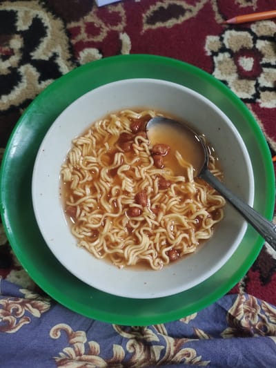 A bowl of instant noodles sits on a green plate, which is on a patterned rug. A spoon is partially submerged in the noodles. The scene is shot from a high angle.