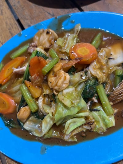 A close-up, eye-level shot showcases a serving of stir-fried vegetables and shrimp on a bright blue plate. The dish, likely prepared in or near Boja, Indonesia, features an assortment of ingredients coated in a glistening brown sauce. Visible components include sliced carrots, cabbage, celery-like stalks, and several plump shrimp. The food is served on a wooden surface, with the texture of the wood grain subtly apparent. The lighting appears to be overhead, casting some reflections on the glossy sauce and the rim of the plate.