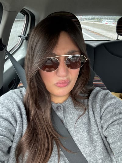 A woman with long, dark brown hair and bangs, wearing a grey knit cardigan and gold-rimmed sunglasses, is seated inside a vehicle in Le Cannet-des-Maures, France. She is secured by a black seatbelt across her chest and looks directly at the camera with her lips pursed in a pouting expression. The background through the rear window shows a wet road surface, with indistinct vehicles visible in the distance, indicating travel on a highway or road. Visible car interior elements include the passenger side rear window frame, a partial view of a black car seat, and the top of a patterned headrest.