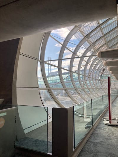 A low-angle, distorted perspective captures the interior of a cylindrical building in Le Mesnil-Amelot, France. The structure appears to be modern, featuring a curved glass facade supported by a white metal framework. The glass panels reveal glimpses of a partly cloudy sky. Sections of the facade are opaque, possibly made of a white material. A grey concrete column is visible in the foreground on the left. Parts of a glass-paneled staircase or walkway with a metal railing and grey floor are visible on the right. A red and white circular fixture is attached to the glass.