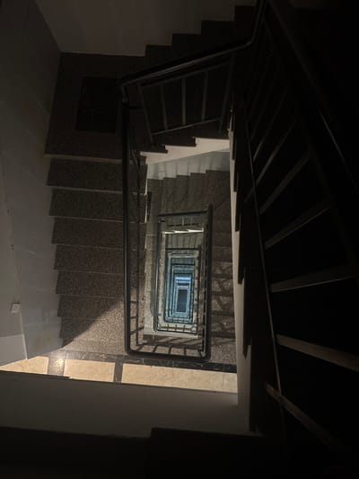 This image depicts a residential stairwell, viewed from an elevated position looking directly down into a multi-story, open central shaft, within Ngũ Hành Sơn Ward, Vietnam. The stairwell consists of square or U-shaped flights of stairs constructed from dark, speckled granite-like material, bordered by black metal railings with horizontal balusters. The scene is dimly lit, with the immediate foreground and sections of the upper right appearing almost entirely dark. Natural or artificial light illuminates the deeper sections of the stairwell, particularly within the central void, creating a repeating pattern of light and shadow on the descending steps and walls. The lowest visible point exhibits a rectangular area of brighter illumination, suggesting a light source or an exit. No individuals or personal effects are present.