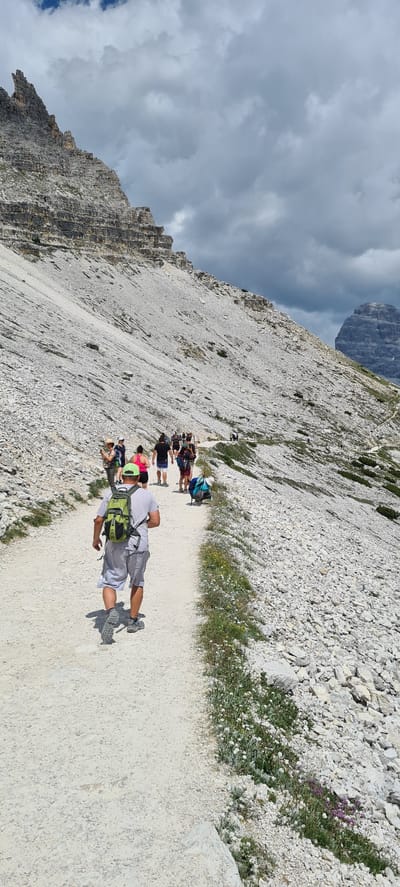 The image depicts a group of people hiking on a gravel path alongside a steep, rocky mountainside. The scene is set in a mountainous region, with a large, imposing mountain visible in the background under a cloudy sky. The path is well-worn and appears to be a popular hiking trail.

The subjects are hikers, predominantly dressed in casual outdoor clothing, including shorts, t-shirts, and backpacks. Some are carrying hiking poles. The hikers are spread out along the path, with a few in the foreground and more further back. The person in the immediate foreground is lying down on the path, possibly resting or posing for the photo, while others are walking or standing. A few individuals are wearing brightly colored shirts, adding a splash of color to the otherwise muted tones of the mountain and path.

The activity taking place is hiking. The setting is a rugged, mountainous landscape, likely in the Dolomites given the location context. The time of day appears to be daytime, given the lighting. The weather is overcast with dramatic clouds in the sky, suggesting a potentially changeable mountain climate. The overall mood appears relaxed, with people enjoying the outdoor activity and scenery.

Notable details include the texture of the rocky terrain, the scattered vegetation with small white and purple flowers along the path, and the sheer scale of the mountainside. The sharp, jagged edges of the mountain in the background and the layers of rock are prominent features. There is no visible text in the image.