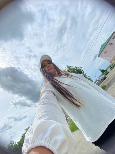 A low-angle, wide-angle shot captures a young woman with long, dark hair wearing a white baseball cap and a white, textured long-sleeved shirt. She is holding the camera out, taking a selfie. The background shows a cloudy sky with dramatic cloud formations, a building with a green roof, power lines, trees, and a road with a car. The location is identified as the city of Akyar, Russia. The woman appears to be outdoors, possibly on a street or pathway.