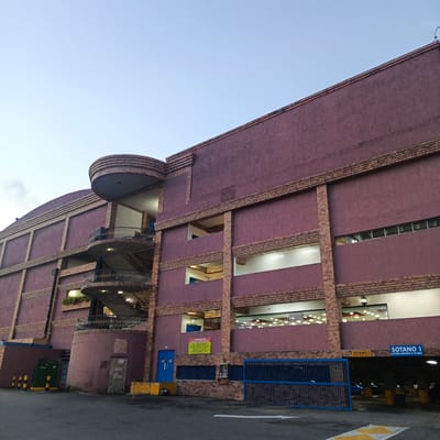 The image shows a multi-level building with a dark purple facade and brick accents. The building has an open-air, circular staircase with black railings on the left side. Various levels of the building have windows and openings revealing interior spaces, some with lights on. A blue metal gate is visible in the foreground, leading into a parking area. Signs indicating "SOTANO 1" (Basement 1) and height restrictions are mounted on the building. The environment appears to be an outdoor urban setting, possibly a commercial or parking structure, in Carrizal, Venezuela, under a pale blue sky. There are no people clearly visible in the image.