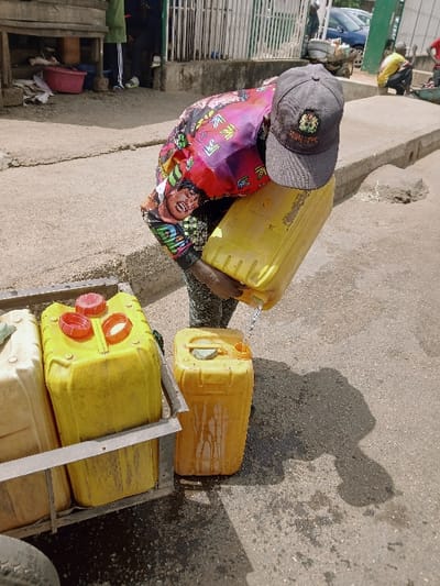 This image captures an outdoor scene in Kadoma, Zimbabwe, on a paved surface under direct daylight. The central figure, wearing a dark grey baseball cap with a distinct emblem and a brightly colored, patterned long-sleeved shirt, is actively pouring liquid from an elevated yellow plastic jerrycan into a similar yellow jerrycan resting on the ground. A continuous stream of liquid is visible during the transfer, and the ground around the lower jerrycan shows signs of spillage, appearing wet.

To the immediate left of the central action, a metal-framed cart or stand holds additional plastic jerrycans; two prominent yellow jerrycans with red caps are visible alongside a lighter-colored, off-white jerrycan. In the background, an urban environment is discernible, featuring a wooden structure or stall, a pink basin, metal fencing, and several parked vehicles, including a blue car. Additional indistinct figures are present in the mid-ground, with one individual in a yellow shirt crouched on the right. Shadows are cast on the paved ground, indicating a bright, sunny day.