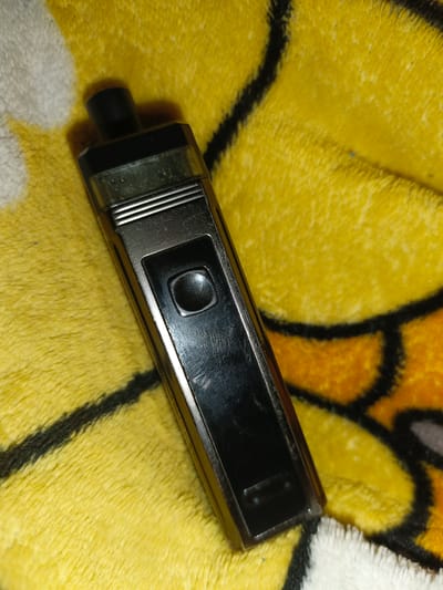 A black, metallic vape pen lies on a yellow and orange patterned fabric. The vape pen is oriented vertically, with the mouthpiece at the top. A black button is visible on the front of the device. The fabric appears to be a soft material, possibly a blanket or towel, with a fuzzy texture. The background is out of focus, suggesting a shallow depth of field. There are no people or other objects clearly visible in the image. There is no information in the image to suggest a location in Shumerlya, Russia.