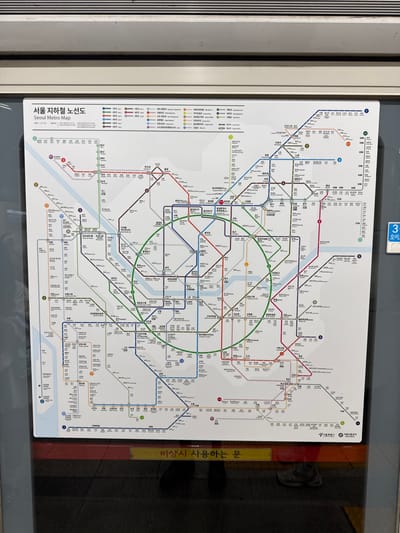 The image displays the interior of a Seoul Metro subway train, likely in South Korea. A large, rectangular "Seoul Metro Map" (서울 지하철 노선도) is centrally mounted on a grey panel, possibly a door or wall. The map, primarily white with multi-colored lines representing subway routes and numerous station names in Korean, occupies the majority of the frame. Below the map, a yellow sign with Korean text, reading "비상시 사용하는 문" (Door for emergency use), is visible. Reflections of the lower legs and feet of at least two individuals are discernible on the dark, reflective surface below the yellow sign, indicating the presence of passengers. Above the map, a horizontally ribbed grey panel is present. To the right, a partial blue sign showing the number "3" and Korean text "오이도" (Oido) is partially visible. The scene depicts a typical operational environment within the Seoul Metropolitan Subway system, which encompasses various districts including 종로구.