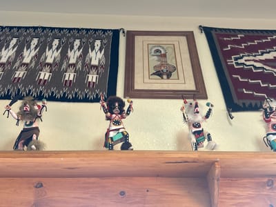 The image shows a wooden shelf with four Kachina dolls displayed on it. Behind the dolls, three woven tapestries hang on a textured wall. To the left, a tapestry features a repeating pattern of stylized figures in gray, white, and red. In the center, a framed piece of art displays a Native American-inspired design with a feathered headdress and pottery. To the right, a tapestry has a geometric zigzag pattern in burgundy, gray, white, and black. The Kachina dolls are diverse in appearance, each adorned with distinct headdresses, clothing, and accessories. They are arranged from left to right on the shelf. The environment appears to be indoors, with the wall painted a light, neutral color. The shelf is made of light-colored wood, showing grain patterns and knots. The overall scene suggests a display of Native American cultural artifacts. Based on the location, this scene is likely situated in Oljato-Monument Valley, United States.