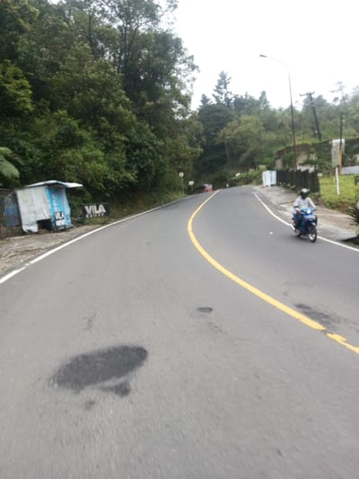 The image displays a two-lane asphalt road traversing a mountainous, heavily vegetated area in Deothang, Bhutan, under an overcast sky. The road features a solid yellow center line and a white right edge line, curving to the right in the distance. Multiple dark, irregular patches, indicative of road damage or repairs, are visible on the asphalt in the foreground. On the left shoulder, a makeshift structure constructed from corrugated metal and white tarp, with blue trim, stands beside a sign displaying "VILA". Dense green vegetation covers the hillside on the left, with a red vehicle partially visible around a distant bend. On the right side, an individual wearing a light shirt, dark trousers, and a helmet rides a blue scooter away from the camera. A street light pole, a utility pole, and several white rectangular objects are situated on the right shoulder, backed by further green foliage and an indistinct white and brown sign.