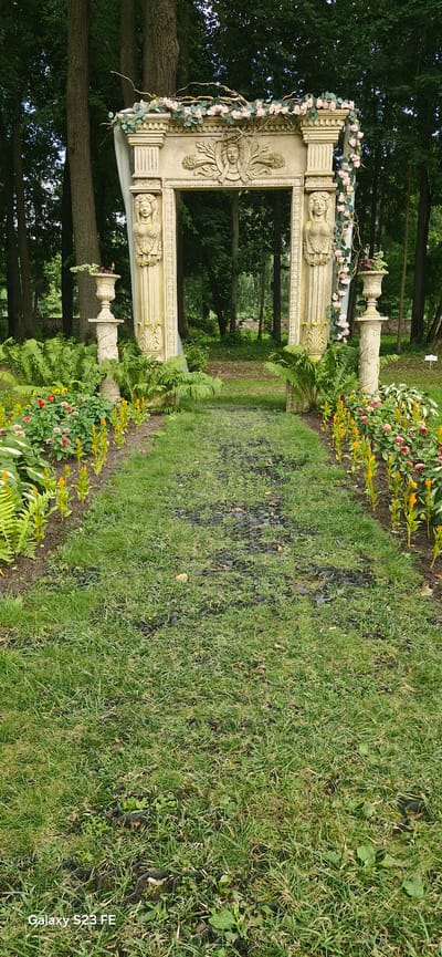 A pathway of green grass leads through a garden towards a stone archway adorned with floral decorations and carved faces. The archway is flanked by stone urns and columns topped with urns. The pathway is lined with flowers and ferns, with a dense forest in the background. The image is a vertical shot, taken in the city of Pakruojis, Lithuania, at the Pakruojis Manor.