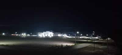 The image depicts a nighttime scene in Meureudu, Indonesia, featuring a large, open field under a dark sky. In the mid-ground, a prominent, brightly illuminated, multi-story building with classical architectural elements, including columns and a central balcony, stands out with its light-colored facade. To the right of this central structure, a row of approximately eight smaller, white, single-story buildings with pitched roofs are also illuminated. Several individuals are observed in small groups near these smaller buildings. To the far left, additional smaller, illuminated structures are visible.

A paved road or pathway curves from the lower right foreground towards the center, bordered by at least three operational streetlights mounted on utility poles. Overhead power lines span the scene. The foreground consists of a vast, flat, unpaved or grassy area, partially lit by the distant lights. The overall environment suggests an institutional or public complex, possibly with temporary facilities, at night.