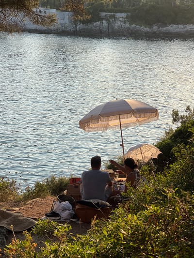 A seaside picnic unfolds with two individuals seated near a body of water. A large, fringed umbrella shades the scene, while a smaller parasol adds a touch of vintage charm. They appear to be unpacking their picnic basket. The sun glistens on the water, creating a serene atmosphere. Lush greenery flanks the picnic spot, enhancing the natural beauty of the setting.