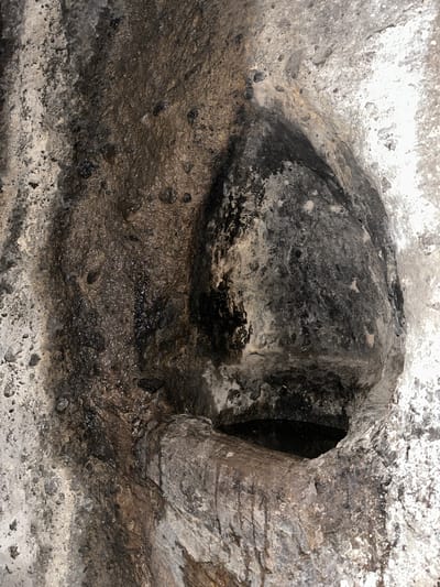This image displays a close-up view of a damp, textured rock formation in Goght, Armenia. The scene is dominated by an irregular, rough stone surface mottled with shades of grey, brown, and black, punctuated by lighter, almost white patches of weathered rock or mineral deposits along the left and right margins. A prominent, dark, tear-drop shaped cavity or recess is visible in the upper-right section of the frame. Below this, a smaller, horizontal opening is present at the bottom-right, partially filled with a pool of dark liquid, consistent with stagnant water. A rough, horizontally oriented stone projection extends from the left into this lower opening, appearing as a natural channel or ledge. The central area of the rock face exhibits numerous glistening wet spots and dark vertical streaks, indicative of active water seepage or previous flow. The surface texture is granular and uneven, with small embedded mineral fragments visible across the damp areas. No people or animals are present.