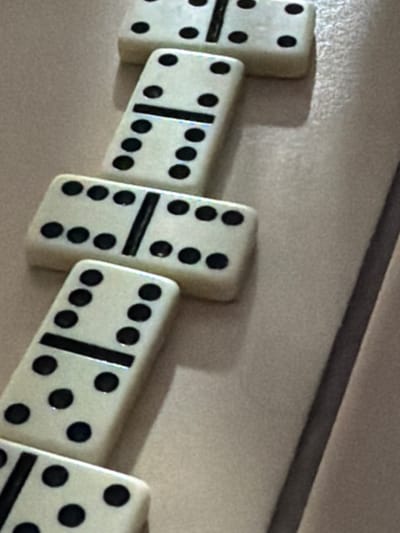This media file shows a close-up, angled view of a series of white dominoes with black dots, laid out diagonally on a textured, light-colored surface. The dominoes are arranged in a chain-like fashion, appearing to fall or be placed in sequence. The first domino on the left is partially visible and shows two dots. The second domino has a vertical line separating its two halves, with three dots on the top half and three dots on the bottom half, totaling a six. The third domino also has a vertical line dividing its halves, with five dots on the top and five dots on the bottom, totaling a ten. The fourth domino shows five dots on its top half and six dots on its bottom half, totaling an eleven. The fifth domino, partially visible on the right, displays five dots on its top half. The lighting suggests an indoor setting, possibly under artificial light, as there are subtle shadows cast by the dominoes on the surface. The background is a neutral, textured material, possibly a wall or a tabletop. There are no people visible, and the focus is solely on the dominoes and their arrangement. The context provided indicates the location is Stryi, Ukraine, but there are no visual cues within the image itself to confirm this specific location. The overall impression is one of a game in progress or a staged arrangement for photographic purposes.