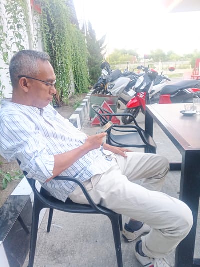In Geulumpangsulu Timu, Indonesia, an adult male wearing glasses, a light-colored striped collared shirt, and light-colored trousers sits on a black plastic chair. He is looking down at a smartphone in his hand. The man is seated outdoors next to a concrete pathway and a low black and white decorative wall. Behind him, several motorbikes are parked in a line. To his right, a dark brown table with a glass and saucer sits on a concrete ground. The environment appears to be a cafe or roadside rest area, with greenery hanging down a wall on the left side of the frame.