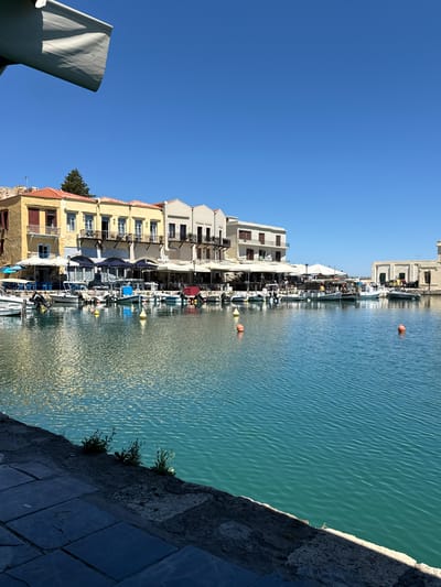 This is a vertical, eye-level, outdoor, daytime photograph of the Rethymno Venetian harbor in Rethymno, Greece. The harbor is filled with clear, turquoise water, with a ripple texture. Several small boats are docked along the waterfront. Behind the boats, a row of colorful, multi-story buildings with awnings and outdoor seating line the harbor. The buildings are light yellow, white, and beige. The sky is a clear, bright blue. In the foreground, a stone walkway and a white awning are visible. The overall scene is serene and picturesque, evoking a sense of Mediterranean charm.