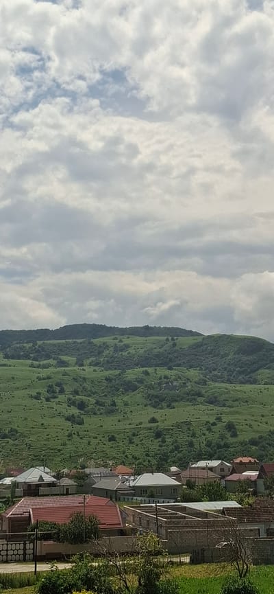 This is a vertical, eye-level shot of a landscape with a foreground of houses and a background of rolling, green hills under a cloudy sky. The houses are predominantly single-family dwellings, some with red tile roofs and others with gray or white ones. One house in the foreground is under construction, with visible brick walls and a concrete foundation. A low stone wall with a patterned top runs along the bottom of the frame, separating the houses from a grassy area. The hills in the background are covered with lush green vegetation and scattered trees, with a darker, tree-covered mountain range forming the horizon. The sky is filled with a mix of white and gray clouds, with patches of blue visible through them. The scene appears to be a rural or semi-rural residential area in Sayukovo, Russia.
