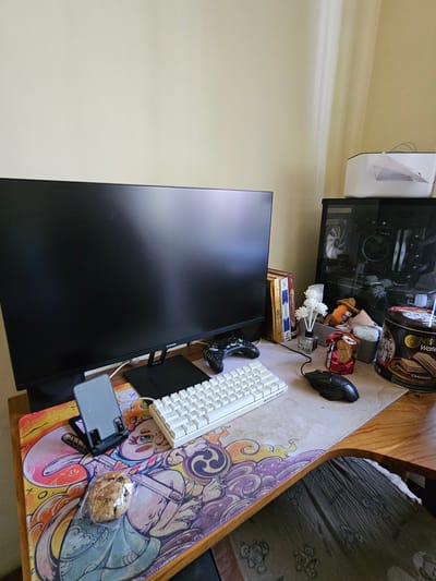 A computer desk is set up in what appears to be a residential setting, possibly in Sidorejo, Indonesia. The desk is positioned in front of a wall and next to a window with natural light casting shadows. On the desk, a large black monitor is prominent, with a white keyboard and a black mouse placed in front of it. To the right of the monitor, a computer tower with a glass side panel is visible, showcasing its internal components and cooling fans. Various decorative items and accessories are scattered across the desk, including a tin of cookies, a small vase with white flowers, a stuffed toy, and a phone stand holding a phone. A game controller rests on the desk to the left of the keyboard. The desk itself is covered by a large, colorful mousepad with an anime-inspired design. A portion of a wooden chair is visible in the foreground, with a textured fabric draped over it. The overall scene suggests a personalized and functional workspace.