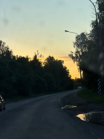 The image shows a rural road at sunset near Pontonny, Russia. The road curves to the right, bordered by dense, dark green trees on both sides. The sky is a gradient of pale blue at the top transitioning to a warm yellow-orange sunset glow near the horizon, with some wispy clouds visible. Streetlights with curved poles are present along the road. In the distance, a white car is driving away from the camera. Closer to the foreground, on the right side of the road, there are two puddles reflecting the sunset sky. A car, partially visible on the left side of the frame, is parked or driving on the road. The overall scene depicts a peaceful, late evening ambiance in a semi-natural environment.