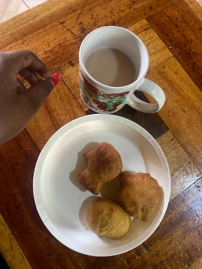 A white plate holds three golden-brown fried dough pastries, while a mug filled with a light-colored beverage rests beside it. The mug features a design with red peppers. A hand with a red nail is reaching towards the plate. The items are set on a wooden surface with a patterned design.