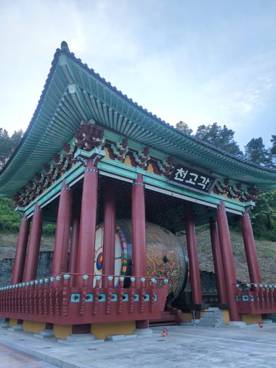A traditional Korean drum pavilion, identified by a black sign with white Korean characters reading "천고각" (Cheongogak), stands in Godang-ri, South Korea. The pavilion features a complex, multi-tiered dark-tiled roof with prominently curved eaves. The underside of the eaves is painted in light blue-green with maroon and white decorative patterns, supported by a layered system of maroon-red brackets with white and blue accents. Eight visible thick, cylindrical pillars, painted deep maroon-red, support the main structure.

A low, ornate railing system, also in maroon-red with light blue-green patterned panels, surrounds the base of the pavilion. The structure rests on a yellow-gold base, which is situated on a light grey concrete or stone platform.

Inside the pavilion, a very large traditional drum is housed. The left visible side of the drum features a concentric circle design with a central red and blue taegeuk symbol, surrounded by colorful rings. The right visible side is adorned with intricate, multi-colored traditional Korean artwork, possibly depicting mythological creatures and clouds.

The pavilion is set against a natural backdrop of a sloping hill covered in green foliage and numerous dark green coniferous trees. The sky above is a pale blue with scattered faint white clouds. The foreground consists of a clean, light grey paved area. Two red fire extinguishers are positioned on the paved ground, one on the left and one on the right side of the pavilion's base. Low grey steps are visible on the right, leading towards the drum entrance. No people are present in the scene.