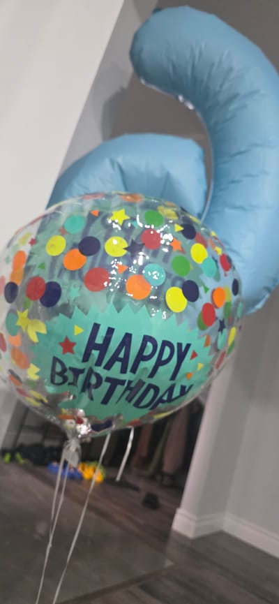 A clear balloon with "Happy Birthday" printed on it is held by two large light blue balloons. The clear balloon is decorated with colorful polka dots and stars. The background is blurry, showing a floor and a wall. The image does not provide any information about the city of Pickering, Canada, or any specific location within it.